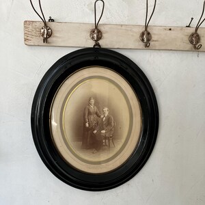 May include: An oval-shaped black frame containing a sepia-toned vintage photograph of two people. The portrait features one person standing and one person seated on a chair. The frame has a gold-colored inner border and a glass front covering the image.