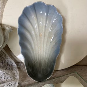 Könnte beinhalten: Eine muschelförmige Keramikschale mit Farbverlauf. Die Schale geht von einem hellen Blau an den Rändern zu einem dunkleren Grau am Boden über. Der Innenraum ist hellcremefarben. Die Schale steht auf einem weißen Teller.