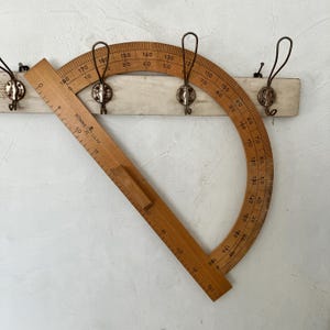 May include: A wooden drafting protractor with a straight edge ruler, featuring black degree markings from 0 to 180 and centimeter measurements up to 50 cm, positioned over a white wall-mounted coat rack with three metal hooks.