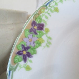 May include: A white ceramic plate with a decorative border of green leaves and purple and light purple flowers. The flowers are stylized and have yellow centers.