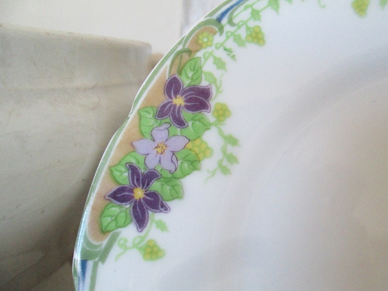 May include: A white ceramic plate with a decorative border of green leaves and purple and light purple flowers. The flowers are stylized and have yellow centers.