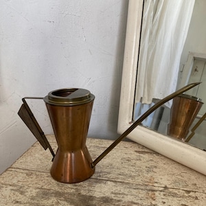 May include: A vintage copper watering can with a long spout and wooden handle. The can has a unique hourglass shape and a brass top. It sits on a weathered wooden surface, with a mirror in the background.