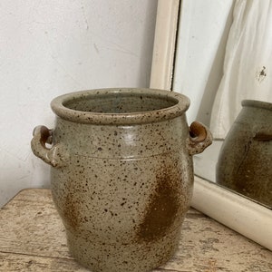 May include: A speckled grey ceramic crock with two handles. The crock has a brown glaze and is slightly rounded.