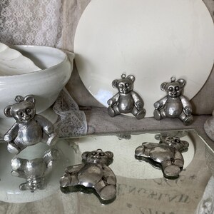 May include: Five silver teddy bear ornaments with a shiny finish. The ornaments are arranged on a white surface and a mirrored surface.
