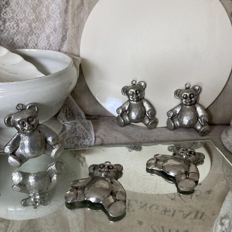 May include: Five silver teddy bear ornaments with a shiny finish. The ornaments are arranged on a white surface and a mirrored surface.
