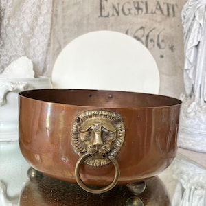 May include: A round copper bowl featuring a prominent brass lion head handle with a ring on the front. The bowl has a smooth, polished finish and is supported by three small, rounded feet at the base, showcasing a metallic construction.