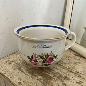 May include: A white ceramic chamber pot featuring a blue rim, a gold-toned edge, and a handle. The front displays a floral pattern of pink roses and green leaves with the script text "À la Mariée" printed in a dark brown colour above the flowers.