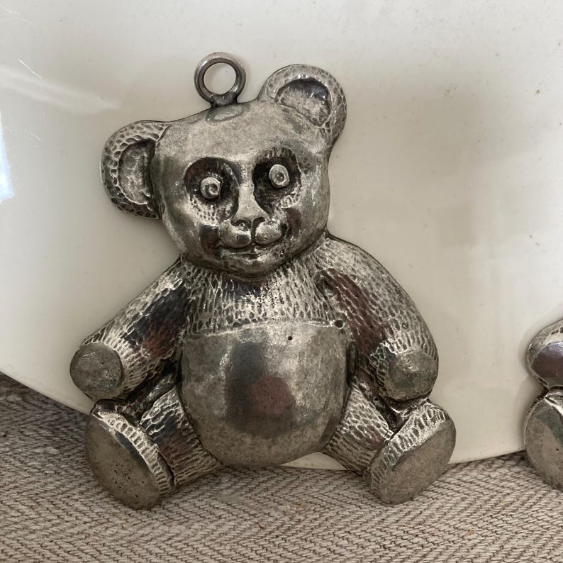 May include: A silver-toned teddy bear charm with a textured surface. The bear has large, round eyes and a small, upturned nose.
