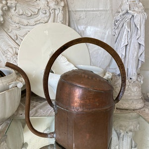 Peut inclure: Un arrosoir en cuivre avec un long bec et une poignée formant un cercle. La carafe a une base hexagonale et un sommet arrondi. La carafe est posée sur une surface réfléchissante.