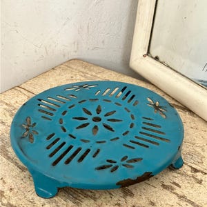 May include: A round, turquoise-colored metal trivet featuring a decorative cut-out pattern of a central flower surrounded by radiating lines and small floral motifs. The piece has three short legs and shows visible signs of wear and rust.