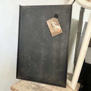 Könnte beinhalten: Ein rechteckiges, dunkelgraues Metalltablett mit einem Used-Look-Finish. Ein Vintage-Foto ist mit einem schwarzen Magneten befestigt. Das Foto zeigt ein junges Kind. Das Tablett liegt auf einer Holzoberfläche.