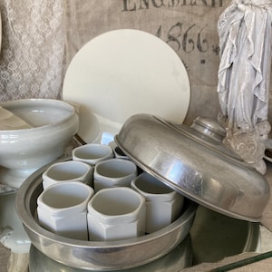 Peut inclure: Un plat de service en métal argenté avec un couvercle et six tasses en céramique blanche à l'intérieur. Le plat est sur une surface miroir.