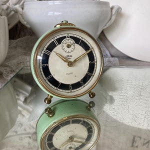 Könnte beinhalten: Ein Vintage-Wecker in Mintgrün und Gold mit einem schwarz-weißen Zifferblatt. Das Zifferblatt hat goldene Zeiger und das Wort "DUETT". Die Uhr hat eine runde Form und steht auf kleinen goldenen Füßen.