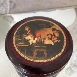 May include: A round wooden box with a dark red finish. The lid features a vintage illustration of children gathered around a table. The illustration is surrounded by a gold border.
