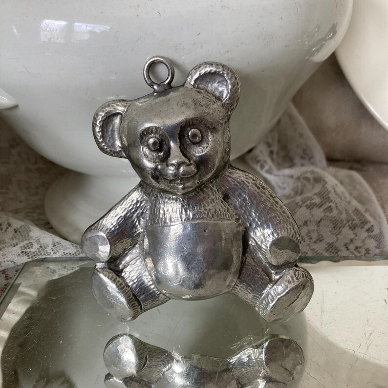 May include: A silver-colored teddy bear charm with a detailed face and textured fur. The charm is a vintage style and would be a great addition to a jewelry collection.