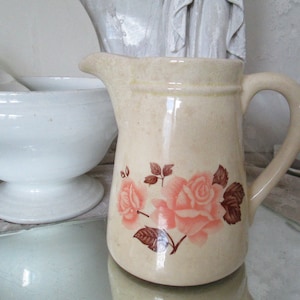 May include: Cream-colored ceramic pitcher with a floral rose design in pink and brown. The pitcher has a handle and a spout. A white bowl is visible in the background.