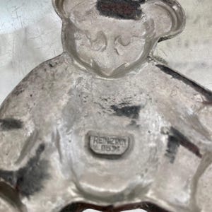 May include: A silver-colored pewter figurine of a bear wearing a hat. The bear is sitting with its legs crossed. The figurine is stamped with the words "REINZINN 95%" on the bottom.