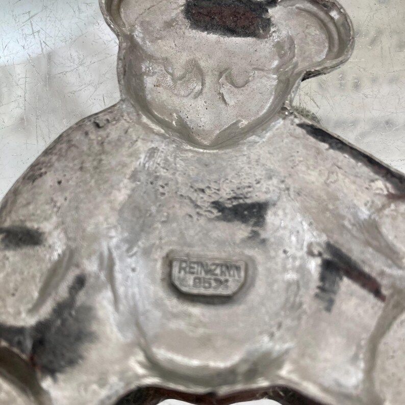 May include: A silver-colored pewter figurine of a bear wearing a hat. The bear is sitting with its legs crossed. The figurine is stamped with the words "REINZINN 95%" on the bottom.