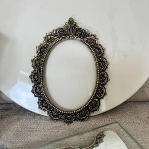 May include: An oval-shaped picture frame with an antique bronze finish. The frame features intricate, raised scrollwork and floral-inspired detailing along the outer edge, with a beaded inner border surrounding the central oval opening.