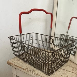 Op de afbeelding: Een rechthoekige draadmand met een rode handgreep. De mand is gemaakt van donker metaal en heeft een rasterpatroon. De handgreep is gebogen en rood geschilderd. De mand staat op een wit, verweerd houten oppervlak.