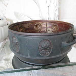 Puede incluir: Un macetero grande, redondo, de metal verde con un diseño decorativo en relieve de rosas y hojas. El macetero tiene un borde dorado y una manija en un lado.