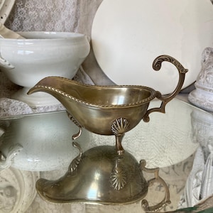 May include: Vintage silver-toned gravy boat with a curved spout and a decorative handle. The boat features a shell-like design on the base and a detailed rim. The gravy boat is a vintage piece, ideal for serving sauces and gravies.