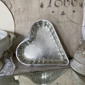 May include: A silver heart-shaped metal mould with a fluted edge. The mould is sitting on a textured surface with the year 1866 printed on it.