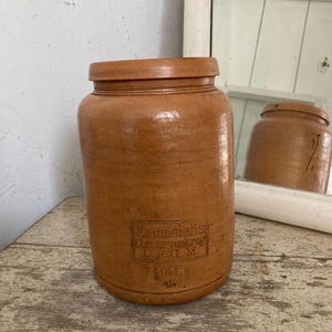 Puede incluir: Un tarro de gres marrón con tapa. El tarro está etiquetado con "Krummeichs Conservenkrug D.R.G.M. 1 1/2 L. 45".