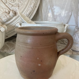 May include: A brown stoneware mug with a handle. The mug has a cylindrical shape with a slightly flared rim and two horizontal lines around the middle. The mug is a warm brown color with some darker spots.