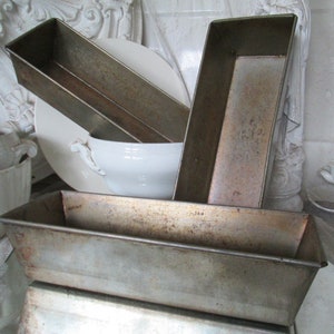 Könnte beinhalten: Drei antike Metall-Brotsformen, zwei sind übereinander gestapelt, die dritte lehnt an dem Stapel. Die Formen sind silberfarben und haben einen abgenutzten, rustikalen Look.