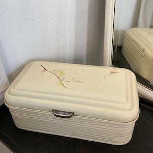 Puede incluir: Panera vintage de color crema con un diseño floral en la tapa. La caja rectangular tiene un diseño acanalado en los laterales y un asa metálica. El diseño floral incluye detalles en rosa, amarillo y verde.