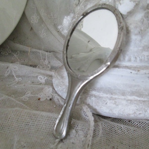 May include: A silver hand mirror with a rounded handle and a clear glass surface. The mirror is lying on a white lace tablecloth.