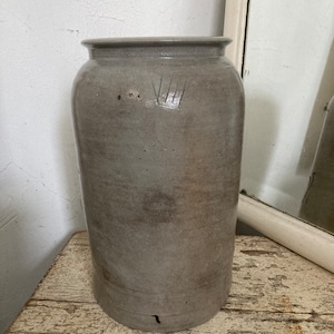 May include: A tall, gray ceramic jar with a wide mouth and a slightly rounded bottom. The jar has a worn, rustic look and has a series of vertical lines etched into the side.