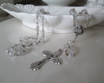 May include: A silver rosary with clear glass beads and a silver crucifix. The rosary is lying on a white surface.