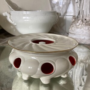 Op de afbeelding: Witte keramische thee-infuser met een gouden rand en rode accenten. De infuser heeft een ronde, open bovenkant en een basis met gaten.