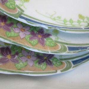 May include: A stack of four white ceramic plates with a floral design. The plates feature a border of green leaves and blue and white stripes. The center of the plates features a design of purple, pink, and yellow flowers.