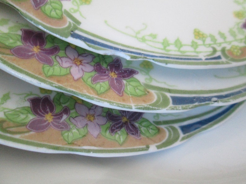 May include: A stack of four white ceramic plates with a floral design. The plates feature a border of green leaves and blue and white stripes. The center of the plates features a design of purple, pink, and yellow flowers.