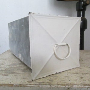 Könnte beinhalten: Eine weiße Metallbox mit einer grauen Metallseite und einem Metallgriff. Die Box hat eine quadratische Form und ein schlichtes Design.