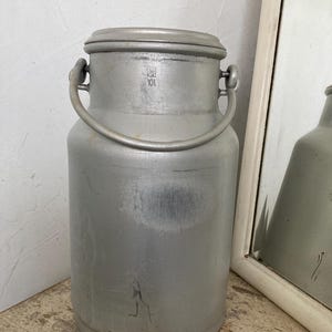 Könnte beinhalten: Ein silberfarbener Milchkrug aus Metall mit Deckel und Griff. Der zylindrische Krug hat ein Fassungsvermögen von 10L, wie auf der Vorderseite angegeben. Er steht auf einer alten Holzoberfläche. Die Höhe beträgt ca. 30 cm.