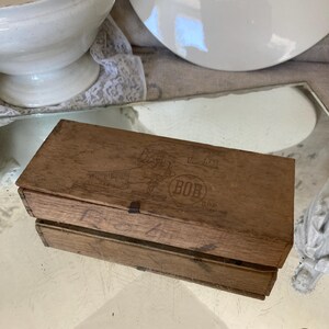 以下が含まれることがあります： 蝶番付きの蓋が付いた木製の箱。箱には「BOB」のラベルが貼られており、ブラシを持った手の絵が描かれている。箱は状態が良く、多少の摩耗がある。