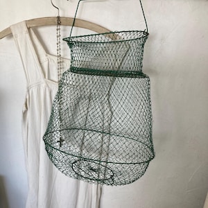 Può includere: Un cestino in rete metallica verde con fondo arrotondato e parte superiore cilindrica. Il cestino è sospeso da un manico in filo di ferro verde. Un capo di abbigliamento bianco è appeso a una gruccia di legno sullo sfondo.