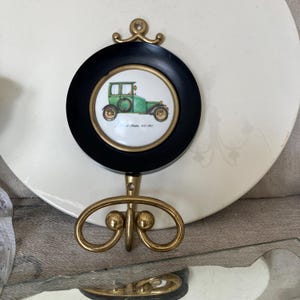 以下が含まれることがあります： 白い背景に、ヴィンテージの緑色の車のイラストが描かれた黒い円形のフレームが特徴の装飾的な壁掛けフック。フックは真鍮仕上げで、装飾的なデザインと上部に小さなループが付いています。