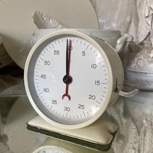 May include: A vintage cream-colored kitchen timer with a white face, black and red hands, and a metal winding lever. The timer sits on a cream-colored base with a green edge. The face displays numbers from 5 to 60.