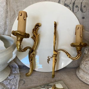 May include: Two antique gold-colored wall sconces with candle-shaped lights. The sconces have ornate, curved arms and decorative leaf accents. The background includes a white plate and a glimpse of a lace-covered surface.