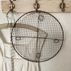 May include: A rusty, round, wire mesh rack with three circular openings hangs from a white wooden rack with hooks. The rack is hanging in front of a white wall.