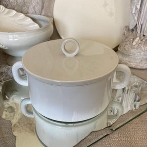 Puede incluir: Un plato de servir de cerámica blanca con tapa y dos asas. La tapa tiene un pomo circular en la parte superior.