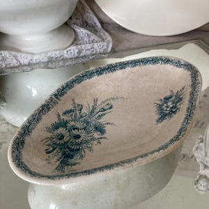May include: An oval-shaped, antique ceramic dish with a cream-colored base and a blue floral design. The dish features a detailed border and two floral bouquets. The dish is likely vintage and adds a touch of elegance to any table setting.
