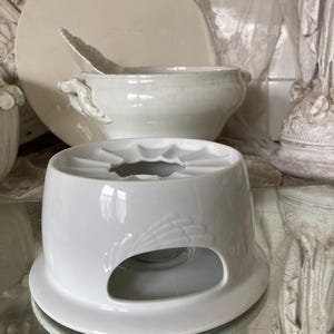 Könnte beinhalten: Weiße Keramik-Fondue-Schale mit dekorativem Design. Die Schale hat einen runden Boden und eine zentrale Öffnung für eine Kerze oder eine Wärmequelle.