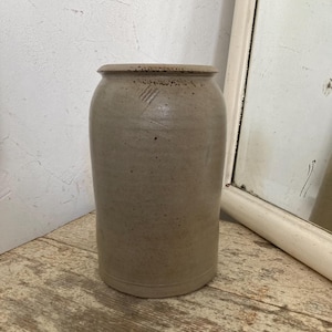 May include: A tall, cylindrical stoneware crock with a slightly flared rim. The antique jar is a muted grey-brown colour with subtle speckling and a few diagonal etched lines near the top. The rustic pottery sits on a distressed wooden surface.
