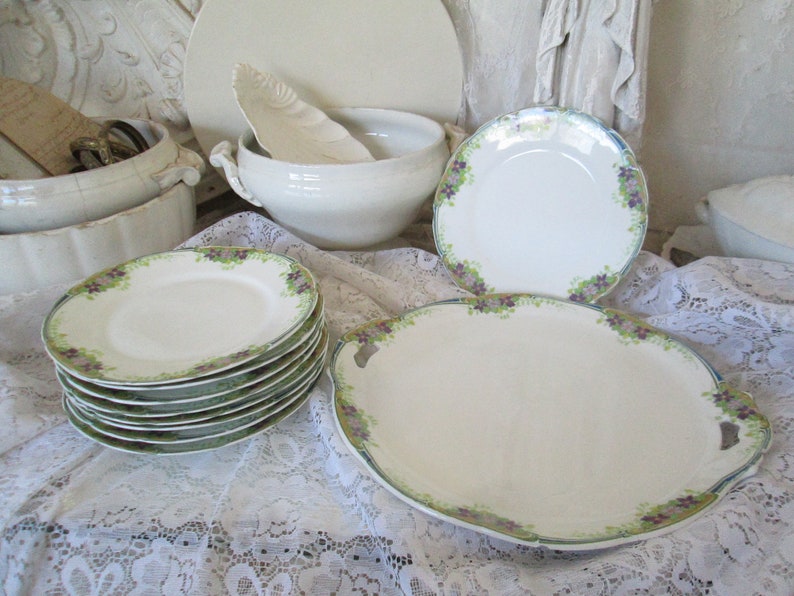 May include: A set of white porcelain plates with a green and purple floral design. The plates are stacked on top of each other and are arranged on a white lace tablecloth.